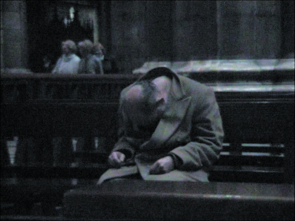 Auf diesem Videostill ist ein älterer Mann mit einer beginnenden Glatze zu sehen, dessen Kinn auf seine Brust gesunken, da er zu schlafen scheint. Er befindet sich dabei auf der Sitzbank eines Kirchensaals.