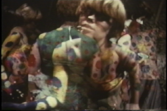 A film still can be seen on which dancing people are depicted with colourfully painted bodies also decorated with dots (polkadots). The bodies seem to blur into each other. 
