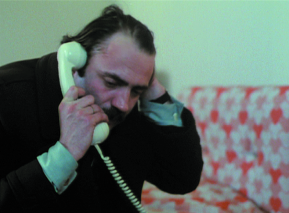 In this video still, a man makes a phone call with a white cord phone while he nervously runs his other hand through his already somewhat light dark hair. Behind it there is a sofa with a white-red flower pattern.