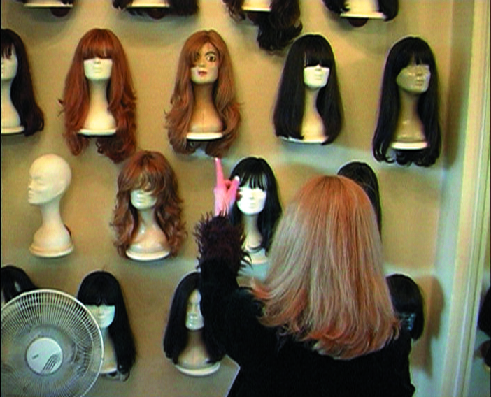 This video still shows the rear view of a woman with long light brown hair standing in front of a shelf and pointing with her left index finger at one of the various wigs on mannequins displayed there.