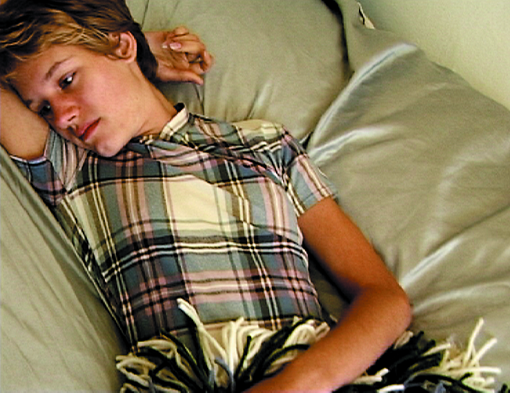 This video still shows a woman with a short haircut, a short-sleeved blouse in light blue tartan and wool skirt, lying dreamily on a bed.