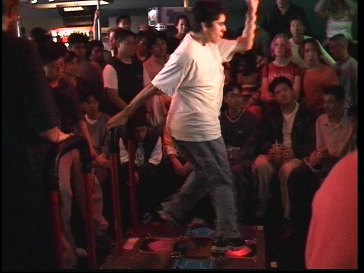 Video Still of a middle-aged woman with a short haircut, wearing jeans and a white T-shirt, dancing on a table in front of many people. Andrea Bowers, Sammlung Goetz Munich
