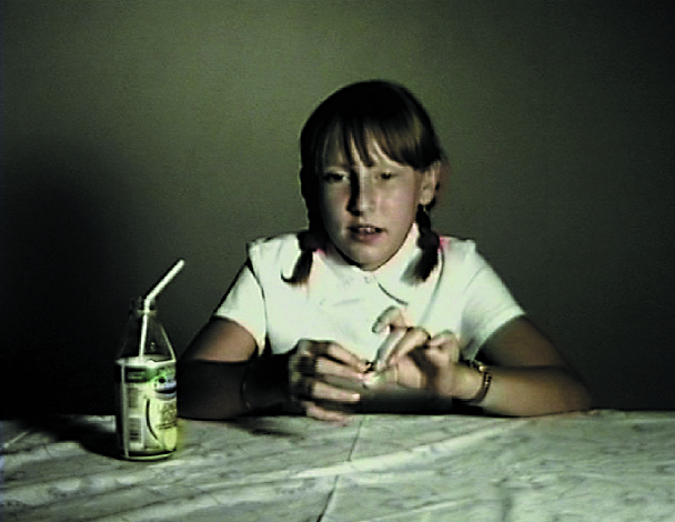Video Still of a girl with two side braids sitting at a table with tablecloth. On it is a small glass bottle with milk and a straw in it. Rosemarie Trockel, Sammlung Goetz, Munich