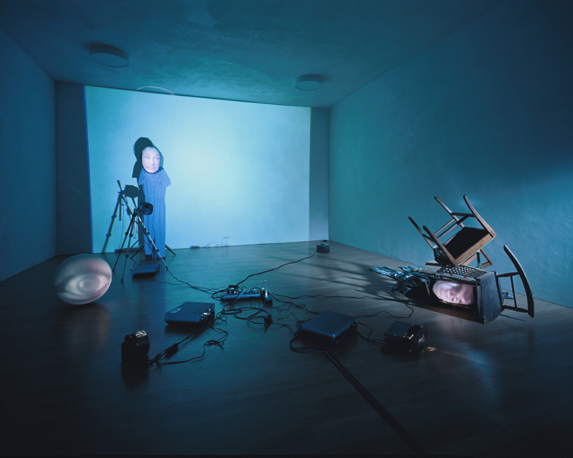 This installation view shows works by the British artist Tony Oursler. Beamers are scattered randomly on the floor, one of them shining a monochrome, bright surface onto the wall. To the left in front of it seems to be a figure whose only plasticity is a contour of the body, projecting a ball with a face on it. There is also a ball on the floor, an eye is projected onto it. Elsewhere there is a tilted, switched on television set with chairs on it. Translated with www.DeepL.com/Translator (free version)