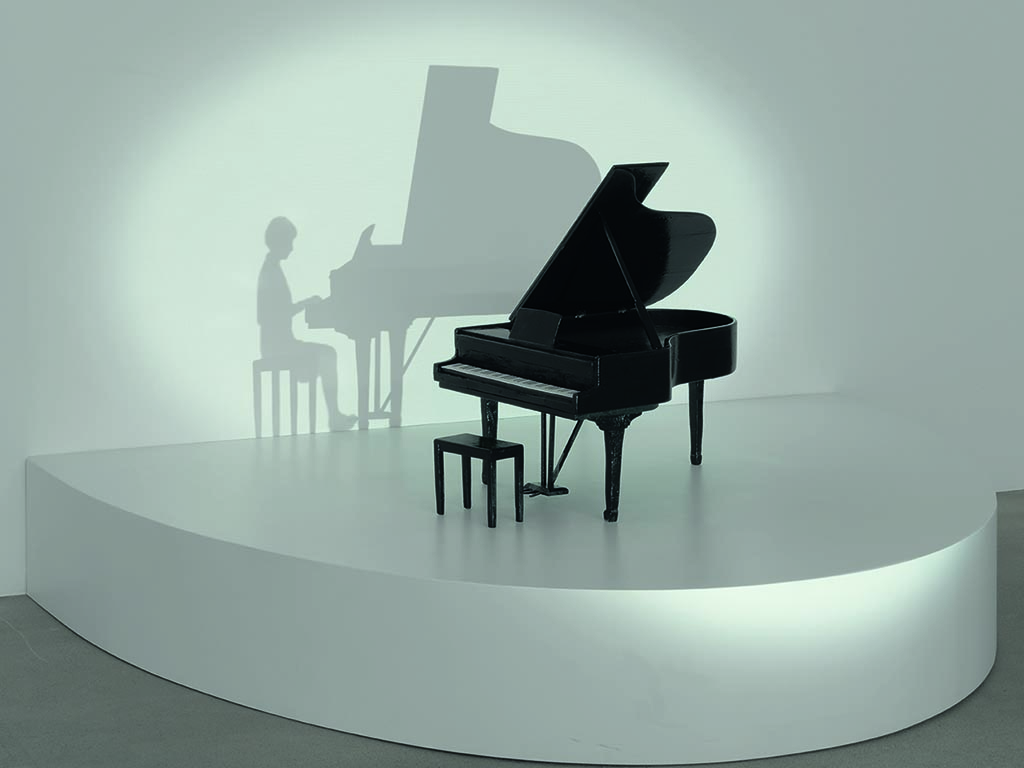 Room installation showing a black grand piano together with a chair on a white, semicircular pedestal. A cone of light seems to fall on the grand piano, casting the shadows of the chair and grand piano, as well as the shadow of a person playing on it, onto the white wall behind it. Zilla Leutenegger, Sammlung Goetz Munich
