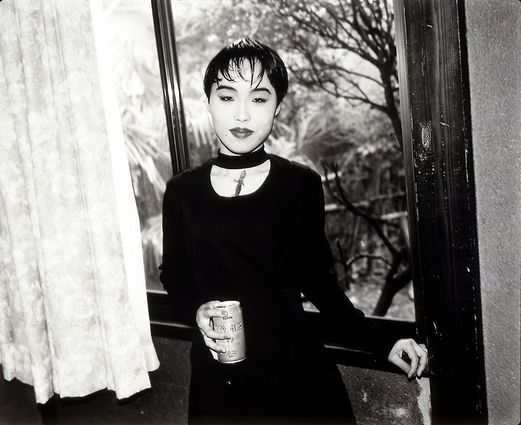 This black and white photograph shows a Japanese woman with short hair at a window of a room. She is holding a can of drink in her hand, wearing a figure-accentuating dress and lots of make-up. In her neckline sits a lizard.