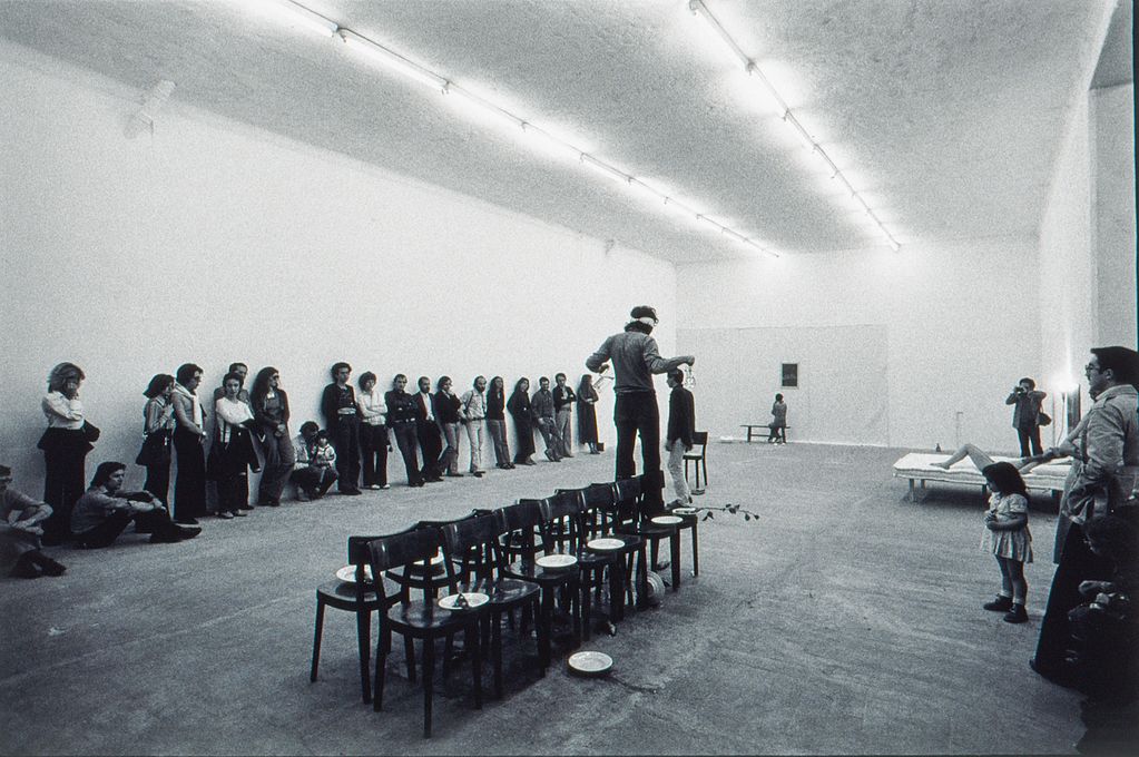 Black and white photograph, a man walks blindfolded on a line of chairs. On some of the seats there are plates, some of them have already fallen on the floor. On the left and right, spectators lean against the walls, on the right, a naked woman lies on a bed. Giorgio Colombo/Pier Paolo Calzolari Sammlung Goetz, Munich