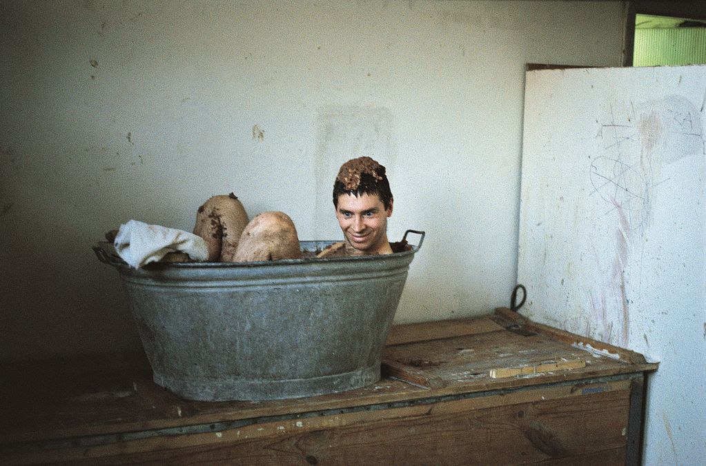 This video still shows the artist Pawel Althamer in an old wash tub on an old big wooden box in a white room. There is a brown mass on his head and he looks funny. Sammlung Goetz Munich