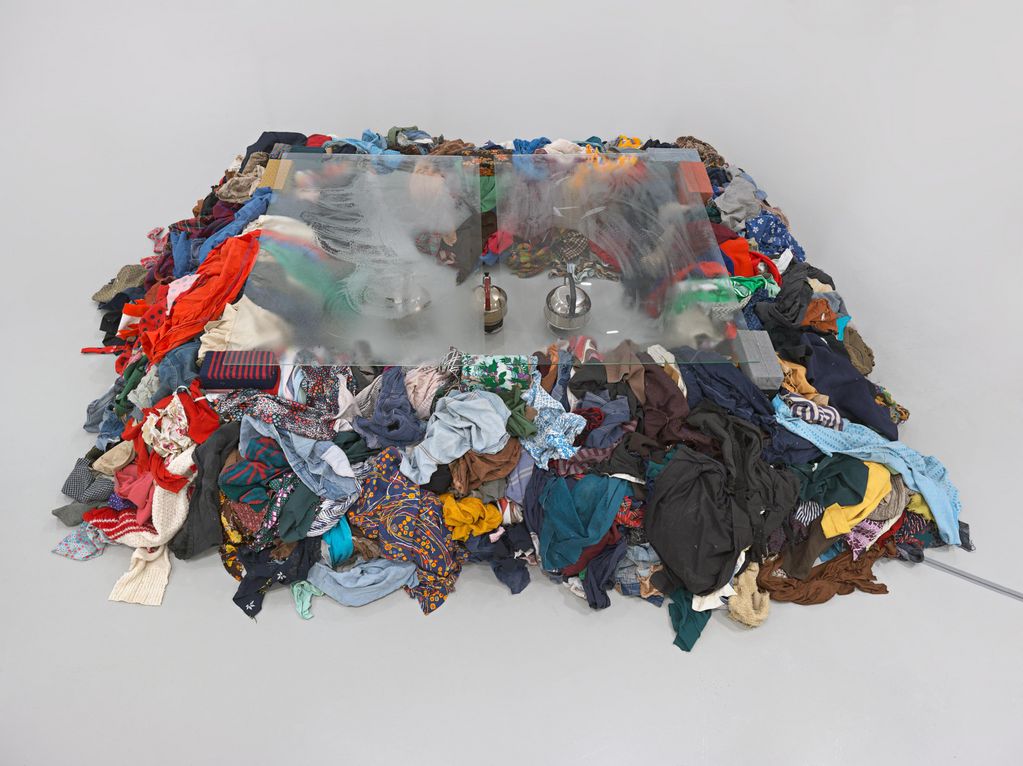 Clothes mountain with two transparent glass plates on top. In the middle of the mountain of clothes there is an open space in which three teapots stand. Michelangelo Pistoletto, Sammlung Goetz Munich