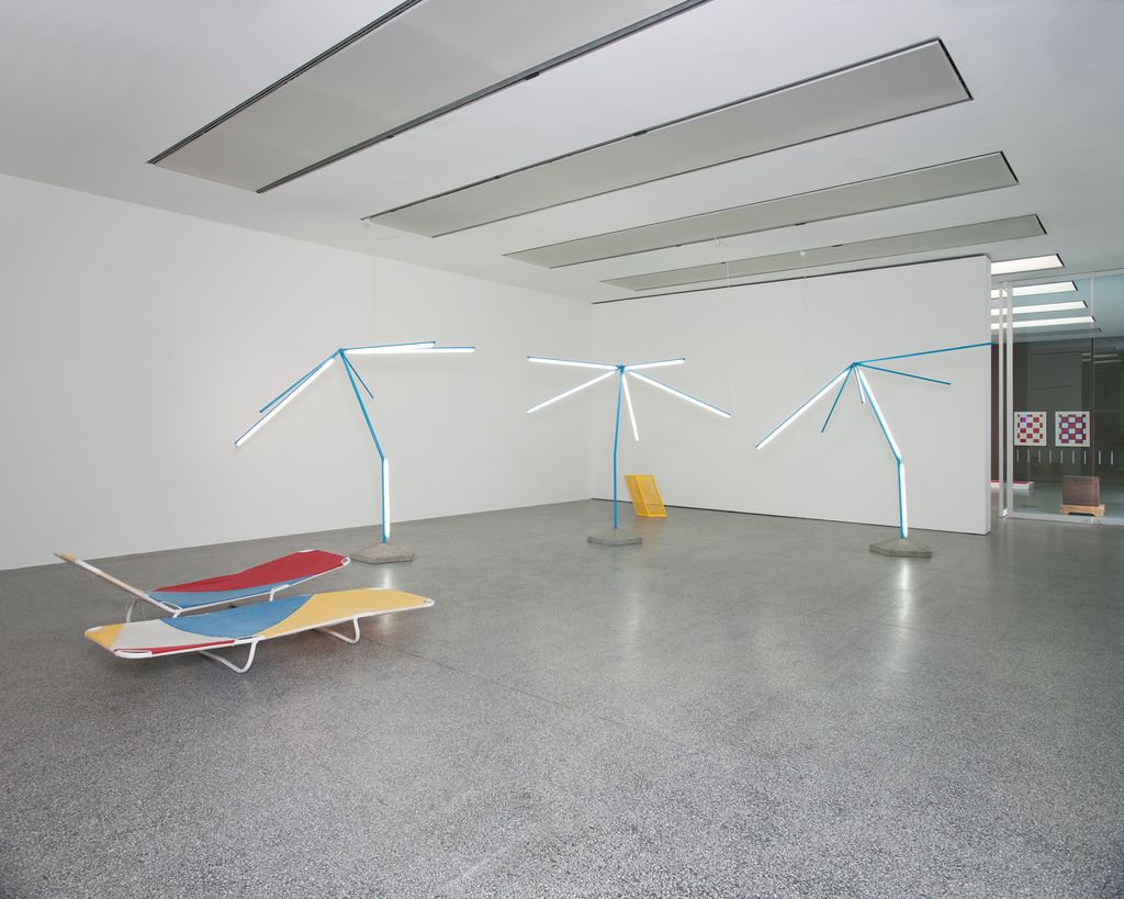 Exhibition view of the work entitled "We are resistant, we dry out in the sun (Our dreams merge and hang in the air like chlorine vapours)" by Martin Boyce. The work consists of two colourful sun loungers and three ribs of parasols with glowing cool white neon tubes attached to their umbrella holders. Martin Boyce, Sammlung Goetz Munich