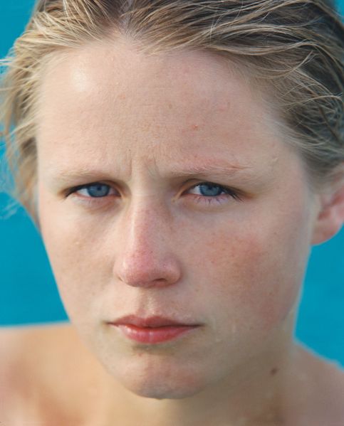Colour photograph, portrait of a young blond, blue-eyed woman. Her shoulders are bare and water is dripping from her face. She is standing in front of a radiant light blue background. Roni Horn, Goetz Collection, Munich  