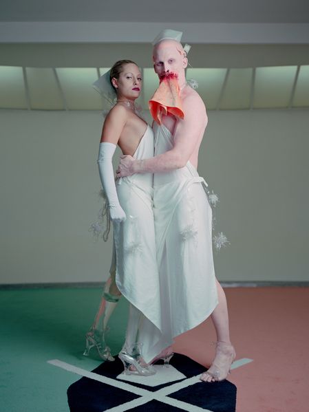 This Production Still shows a woman and a man with white aprons and caps, she with transparent prosthetic legs, he with a kind of bloody cloth in the mouth area. The man holds the woman in his arms, both look directly into the camera and stand on a carpet with a kind of emblem on it.  