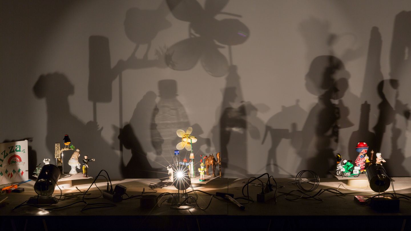 Installationsaufnahme der Arbeiten "Schatten" des Künstlers Hans-Peter Feldmann. Dabei handelt es sich um eine Arbeit, bestehend aus verschiedenen kleineren Artefakten auf einem Tisch, die von mehreren kleinen Lichtquellen angeleuchtet werden und so einen Schatten auf die dahinter liegende Wand werfen. Hans-Peter Feldmann, Sammlung Goetz München
