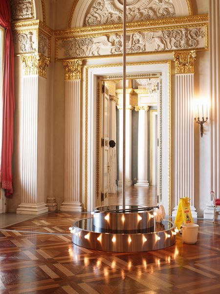 Installation view of the work "Go Go Go!" in an opera hall with wooden floor, pilasters on a door arch and abundant stucco, which is partially decorated with gold leaf. The work itself consists of a pedestal + pole of a gogo dancer, on the pedestal is a mop, next to it is a cleaning bucket and a yellow warning sign. Michael Elmgreen/Ingar Dragset, Sammlung Goetz Munich