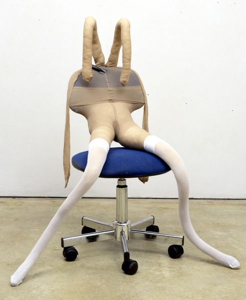 This picture shows a sculpture of stuffed tights in the shape of a human lower body, sitting on a blue upholstered office chair. The shape of the lower body is extended by another stuffed pantyhose, which looks like rabbit ears and is attached to the backrest with a ferrule.