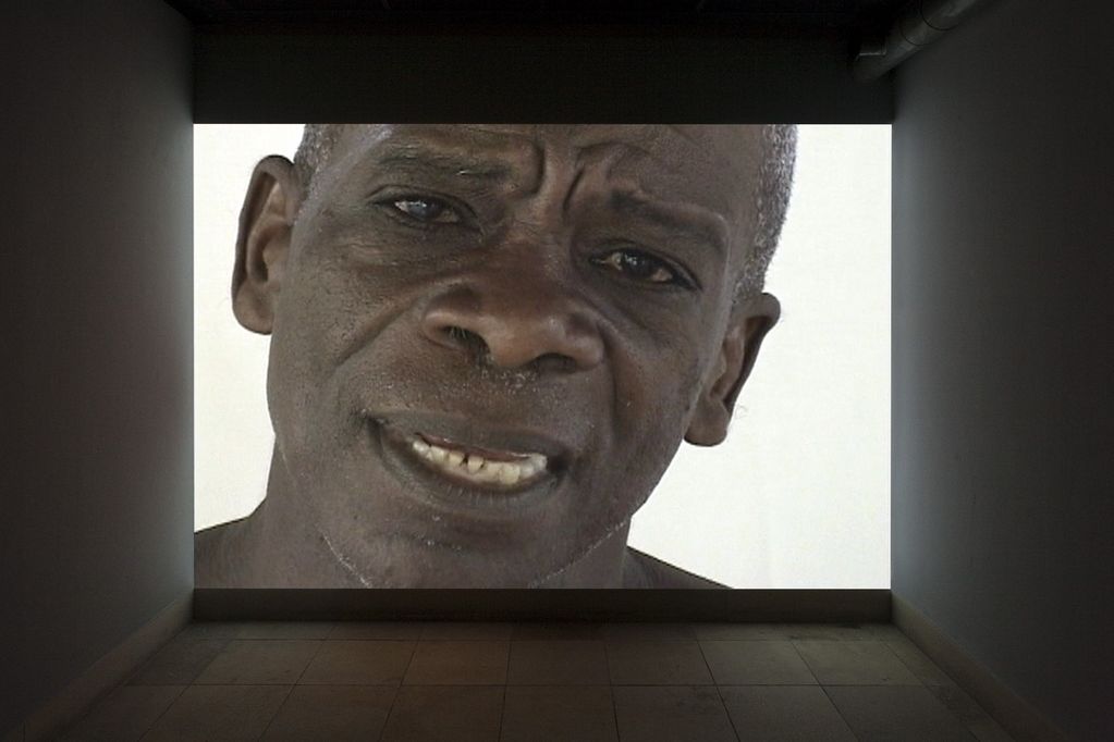 Photography of a dark room with video projection, whose video still is currently showing the face of an elderly man of color. Manuel Echavarrìa, Sammlung Goetz Munich