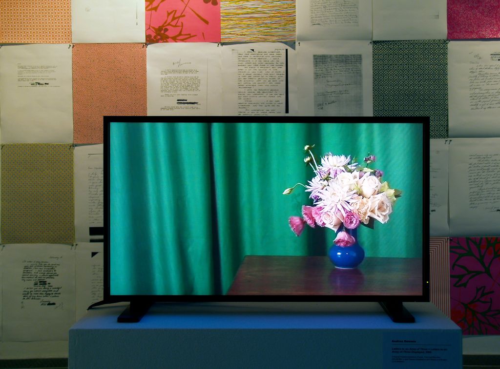 Detail of the installation of the works "Letters to an Army of Three + Letters to the Army of Three Displayed". The monitor shows a still life with a green curtain, wooden table and a vase filled with pink flowers. Behind the monitor, DinA4 papers are hung on the wall, which contain texts or elaborate patterns. Andrea Bowers, Sammlung Goetz Munich 