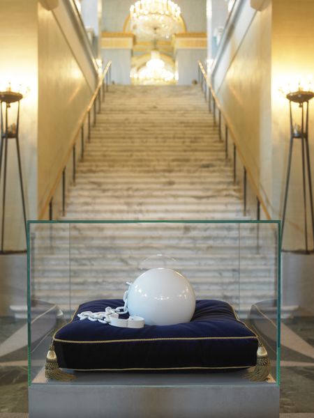 Anklet with ball made of glass in white lying on a blue velvet cushion in a free-standing display case with a base. The display case is located in front of a staircase leading to an opera hall. Michael Elmgreen/Ingar Dragset, Sammlung Goetz Munich