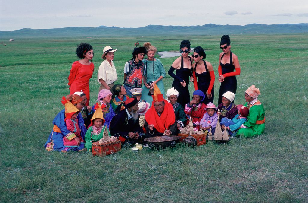 Photography of a group of people standing or sitting in a green landscape with green mountains in the background. On the ground are people with Mongolian features who seem to be preparing a ceremony, while mainly Western women stand behind them and look on curiously. Ulrike Ottinger, Sammlung Goetz Munich