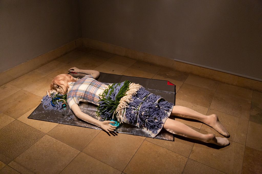 Die Abbildung zeigt eine lebensechte weibliche Puppe mit blonder Perücke, karierter Bluse und Wollrock auf dem Boden auf einer silbernen Decke liegend. Rosemarie Trockel, Sammlung Goetz München