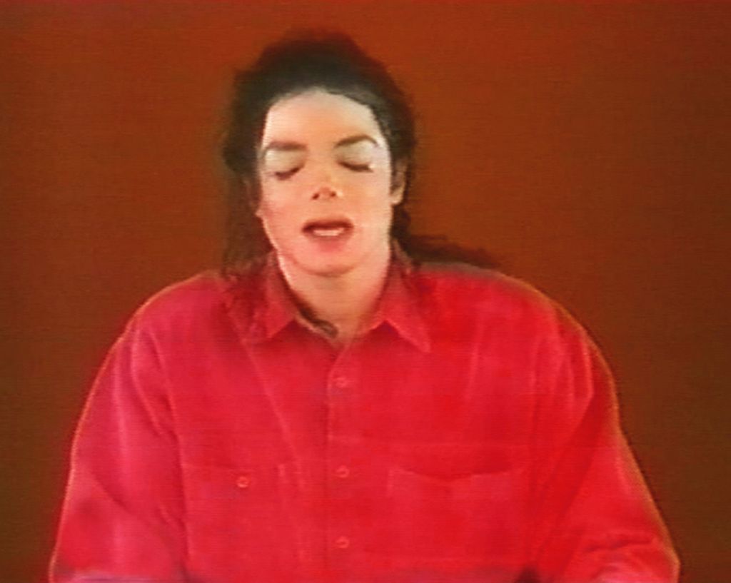 Video Still showing the late pop singer Michael Jackson, with black hair, bright white skin and a red shirt. The singer has his eyes closed and seems to be just in the flow of speech. Paul Pfeiffer, Sammlung Goetz Munich