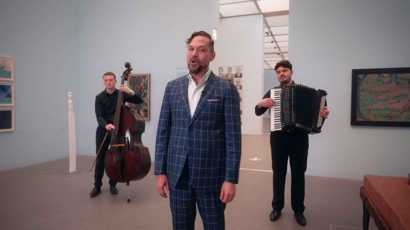 Ein Mann im karierten Anzug steht in einem Ausstellungssaal. Hinter ihm stehen ein Mann mit Kontrabass und einer mit Akkordeon