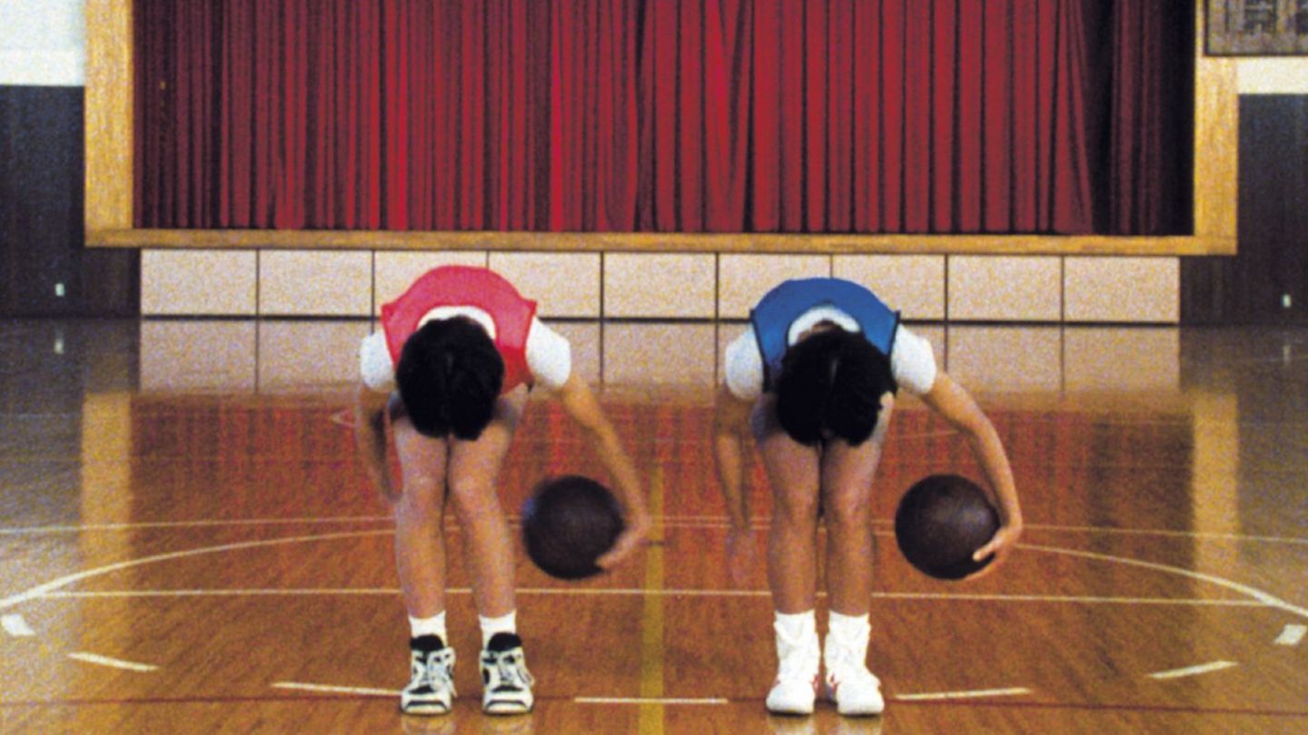 Dieses weitere Filmstill der Arbeit "Goshogaoka" von Sharon Lockhart zeigt die Frontalansicht zwei Basketballspieler(*innen), der eine in einem roten, der andere in einem blauen Trikot den gleichen Trick in gleicher Haltung ausführend in einer Schulturnhalle mit einer Theaterbühne samt zugezogenen Vorhang im Hintergrund.