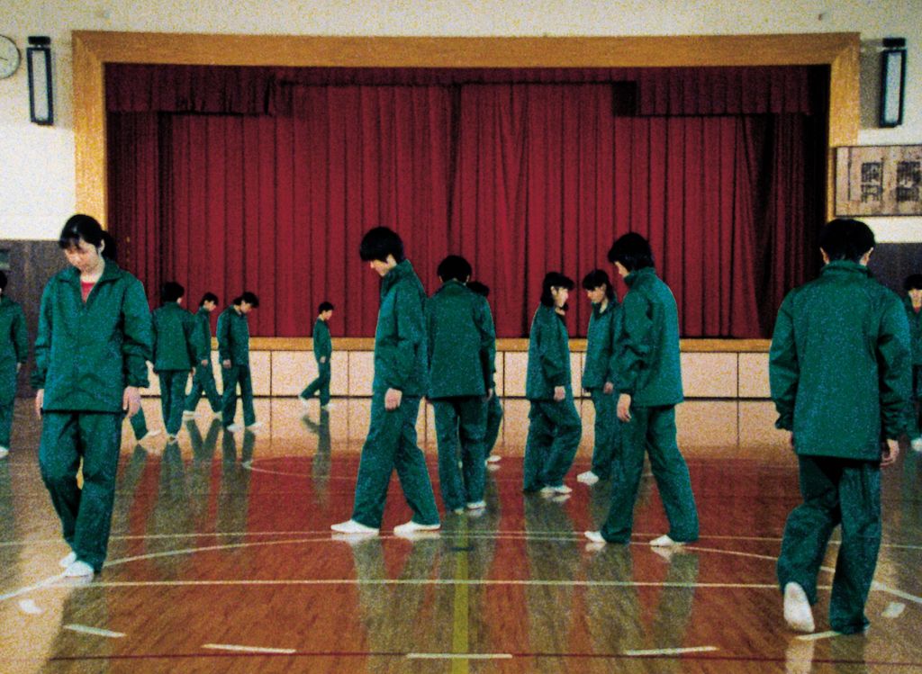 In this film still, you can see Asian-looking pupils in green sports uniforms moving around the basketball court in a school gymnasium. Most of them have their eyes on the floor and seem to follow the lines of the recorded field. In the background, a theatre stage can be seen with its burgundy red curtain drawn.