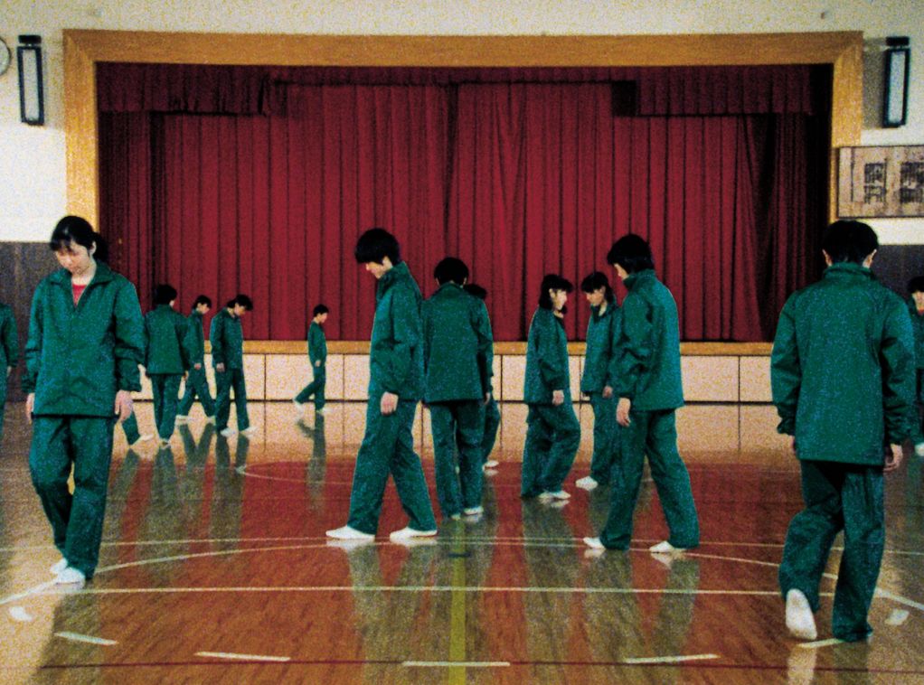 In diesem Filmstill erkennt man asiatisch aussehende Schüler*innen  in grüner Sportuniform, die sich auf dem Basketballfeld in einer Schulturnhalle bewegen. Die Meisten haben den Blick auf den Boden gerichtet und scheinen die Linien des aufgezeichneten Feldes nachzugehen. Im Hintergrund ist eine Bühne zu sehen, deren bordeauxroter Vorhang zugezogen ist.