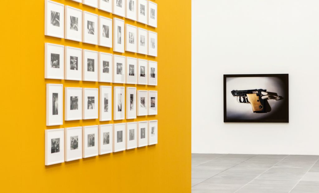 Ausstellungsansicht mit einer mehrteiligen schwarzweißen Fotoarbeit auf einer gelben Wand und einer farbigen Fotografie eines kleinen Revolvers auf einer weißen Wand. Laurie Simmons, Sammlung Goetz München