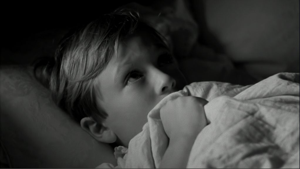 Video Still in black and white showing a close-up of a little boy lying in bed who seems to be holding the bedspread over his mouth in fright. Clement Page, Sammlung Goetz Munich