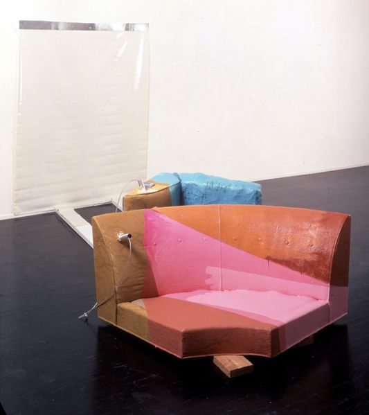 This installation consists of the corner part of a couch, further couch cushions, iron parts, wooden planks and a two-dimensional object on the wall, which was fixed with adhesive tape.  