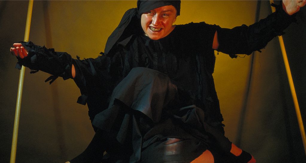 Die Fotografie zeigt eine unangenehm lächelnde Frau ganz in schwarz gekleidet und sitzend. Sie hat ihre Arme erhoben und ihr rechtes Bein über das linke gelegt.