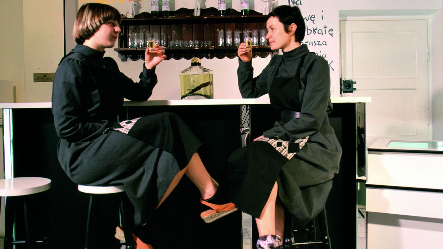 This photograph shows the artists Lucy McKenzie (left) and Paulina Olowska (right) sitting at a bar with filled shot glasses, which they have raised to toast. On the wall behind them are words in Polish, presumably drinks, with a few sheets of paper from their collaborative series "Nova Popularna" hanging between them. Lucy McKenzie/Paulina Olowska, Sammlung Goetz Munich