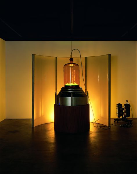 This photograph shows the exhibition view of the work "Kandor 7" by Mike Kelley. In the background, a semicircular curved screen is illuminated by a yellow light source; in front of it is a futuristic sculpture containing a luminous model city under a glass dome. Mike Kelley, Sammlung Goetz Munich