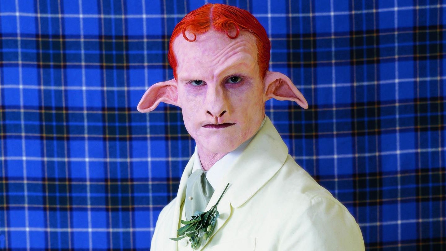 On this production still you can see a Satyr with red hair and a white suit. He stands in front of a tartan fabric in blue.