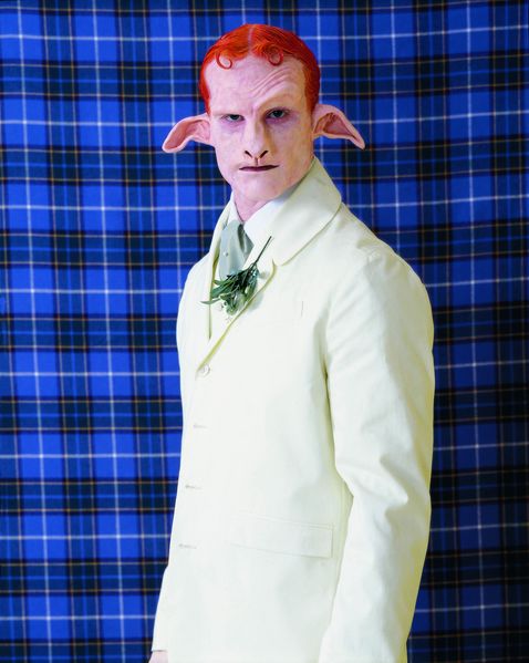 On this production still you can see a Satyr with red hair and a white suit. He stands in front of a tartan fabric in blue.