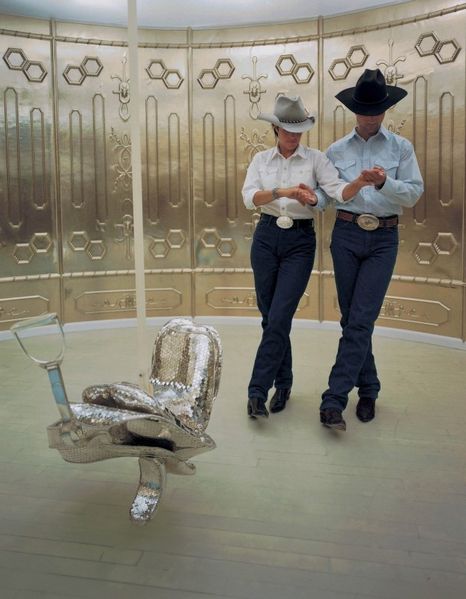 Hier sind ein Mann und eine Frau zu erkennen, die synchron in Cowboykleidung miteinander tanzen. Sie befinden sich in einem Gold vertäfelten Raum, in dem ein mit Pailletten überzogener Pferdesattel hängt.