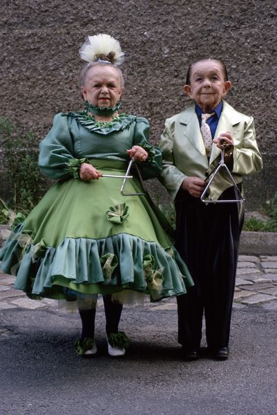 Photograph of two women of small stature in green costumes. One lady is dressed in a green dress with petticoat, ruff and headdress, while the other woman is wearing a tailcoat including tie. They are each holding a triangle in their hands and looking at the camera. Ulrike Ottinger, Sammlung Goetz Munich