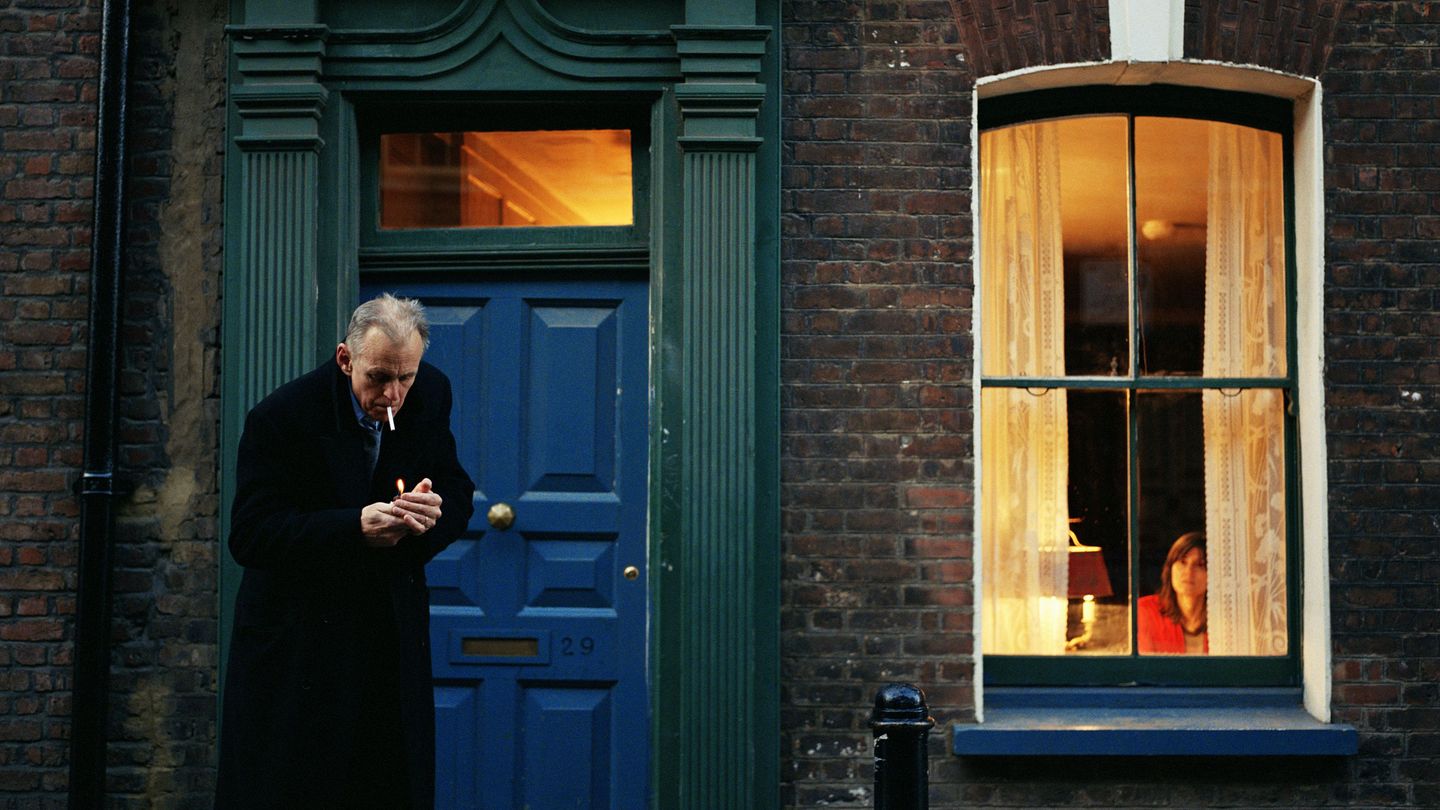 Video Still eines älteren Mannes in dunklem Mantel vor einer Haustür eines Backsteingebäudes, der im Begriff ist, sich eine Zigarette anzuzünden. Sam Taylor-Johnson, Sammlung Goetz München