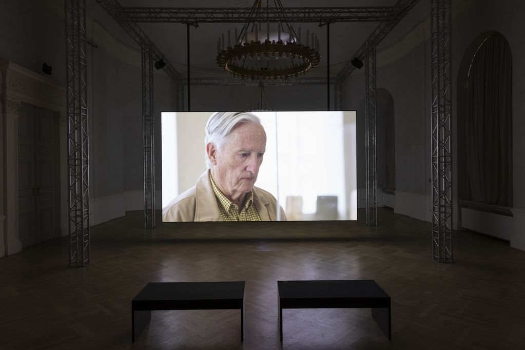 Exhibition space with a screen showing an elderly white-haired gentleman. Teresa Hubbard/Alexander Birchler, Sammlung Goetz Munich