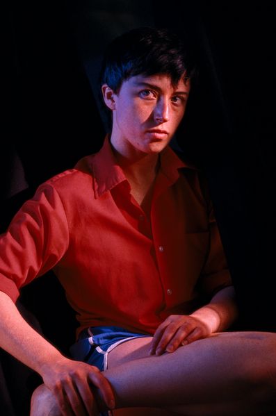Self-portrait of the artist as an androgynous person with short black hair, red shirt and short sports shorts in light blue. The person is sitting and has one foot drawn up to her thigh with one hand. Cindy Sherman, Sammlung Goetz Munich 