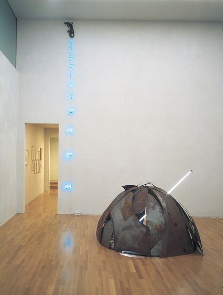 The recording shows an installation view of two works by the artist Mario Merz. At the top of the wall is a preparated caiman, below it is the Fibonacci number series from one to 114 in light blue fluorescent tube charters. On the wooden floor is an igloo made of steel plates of different sizes with a neon tube placed diagonally through its centre.