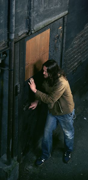 The photograph by artist Jeff Wall shows a long-haired and bearded man who is about to open a door in a dark alley. He seems to be listening and above all cautious. The photograph seems to have been taken from the perspective of an upper apartment on the opposite side.