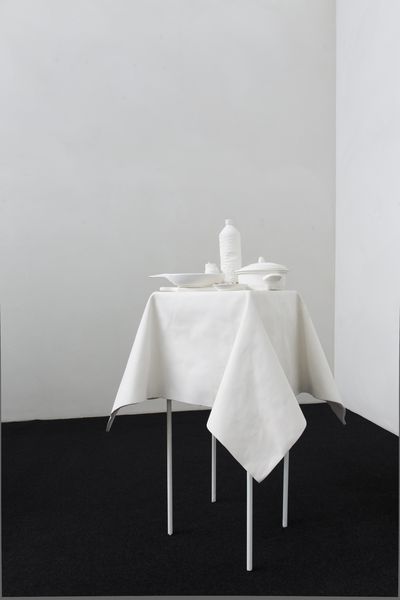 Ein weißer Gipstisch mit langen, dünnen Tischbeinen steht auf schwarzem Grund in der rechten, unteren Bildmitte. Der Tisch selbst ist mit einer Tischdecke bedeckt, die gleichmäßige Falten über die Tischkanten wirft, darauf befinden sich eine Flasche, ein kleiner Teller, ein Topf mit Deckel und ein großer, tiefer Teller. Das komplette Modell besteht aus weißem Gips, der Hintergrund ist weiß gestrichen. Hans Op de Beeck, Sammlung Goetz München