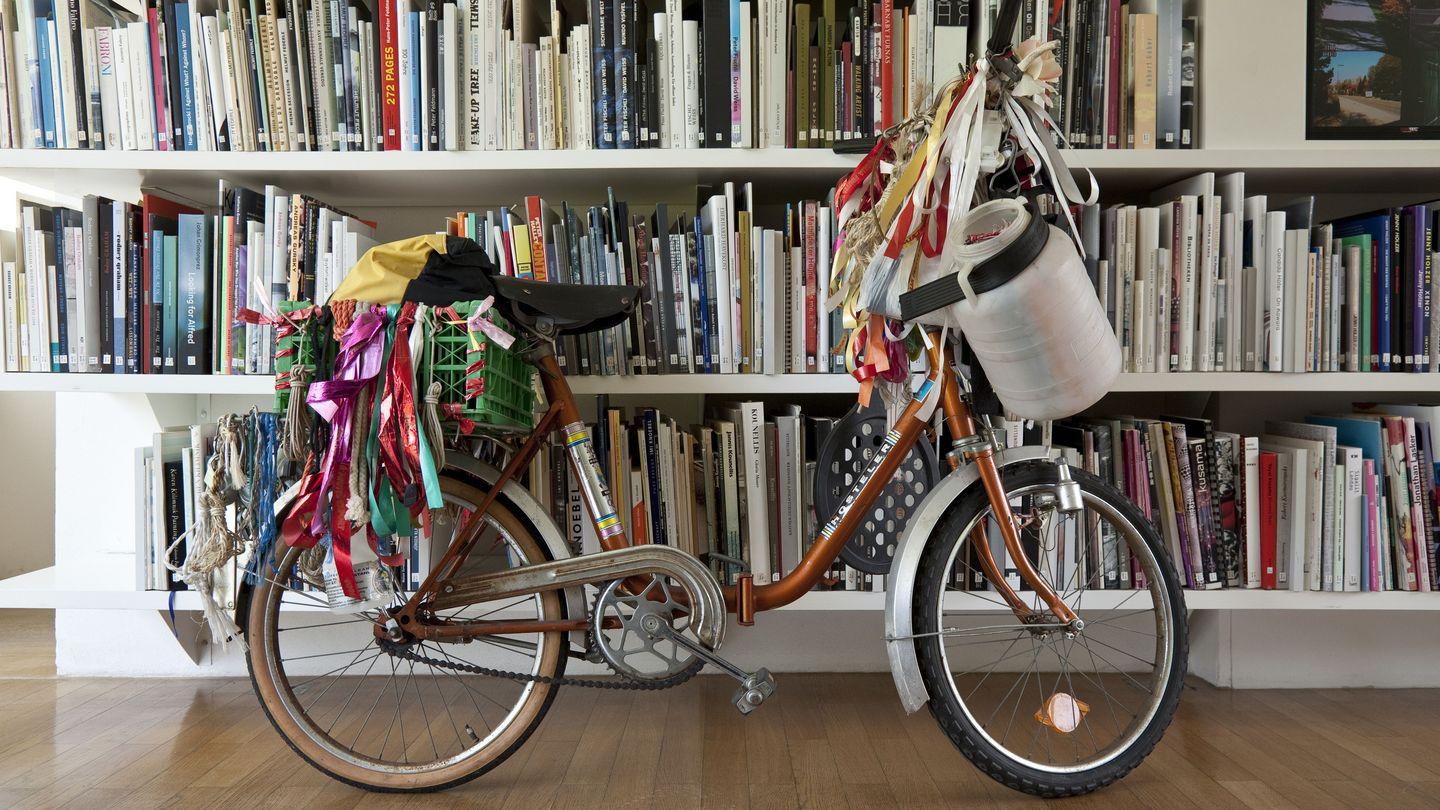 Copper colored folding bike hung with colorful fabric ribbons, a canister, and a toy bat. A green plastic box is attached to the back rack. The bicycle stands in front of a bookshelf, which seems to have been sorted without any order. Andreas Slominski, Sammlung Goetz Munich