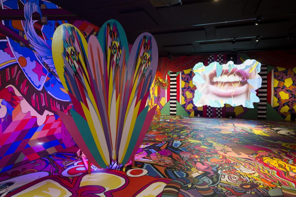 Colourfully designed walls of an exhibition space with a multi-coloured partition wall and a video projection in the middle of one of the colourful walls showing an open mouth. assume vivid astro focus, Sammlung Goetz Munich