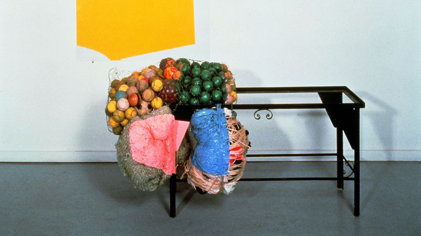 This installation shot contains a two to three dimensional work by Jessica Stockholder. It consists of an acrylic painting in yellow on glass, a black iron frame of a small table and colorful balls in rabbit wire, as well as wrapped sheep wool and other wrapped textiles tied to the table frame. 