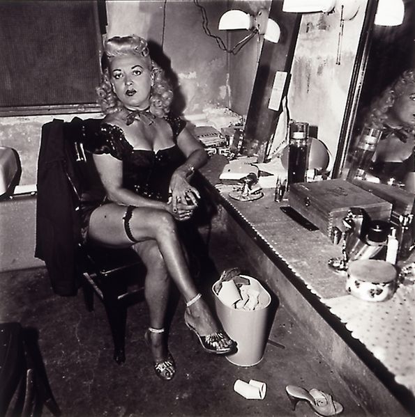 Die Schwarzweißfotografie der ameriikanischen Künstlerin Diane Arbus zeigt eine Burlesque-Tänzerin an ihrem Schminktisch sitzend. Sie hat blonde Haare, ist schon geschminkt, trägt ein knappes Outfit mit Strumpfband, hat die Beine überschlagen und blickt den Betrachter oder die Betrachterin herausfordernd an.