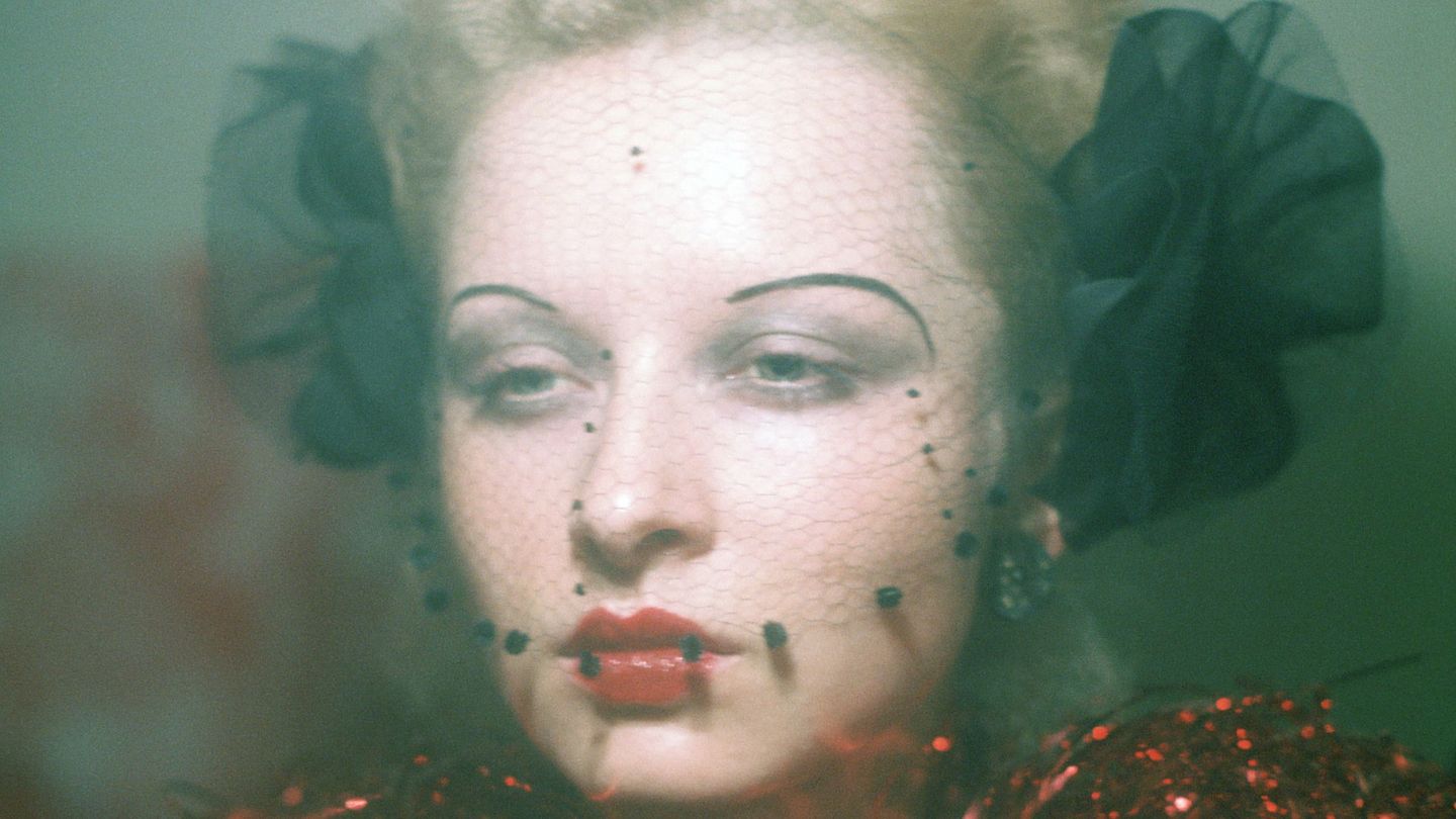 Porträt einer jungen, blonden Frau mit 20er Jahre Make-up, einem schwarzem Netz vor dem Gesicht und einer Halskrause aus rotem Lametta. Ulrike Ottinger, Sammlung Goetz München